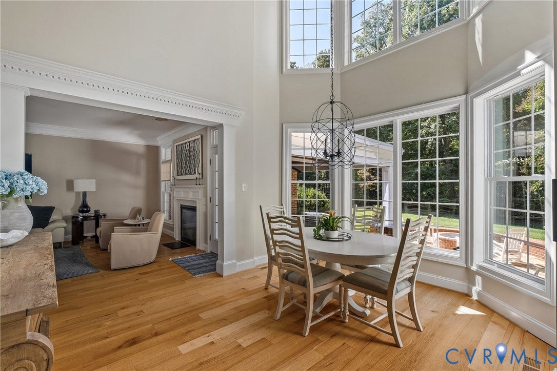 Hardwood flooring, two-story ceiling, decorative chandelier, large windows with exterior landscaping, and an adjacent living area with a fireplace