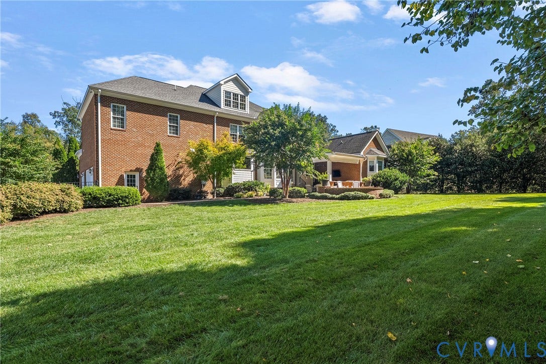 Expansive lawn area with mature perimeter landscaping, brick exterior, and a covered patio with a stone base