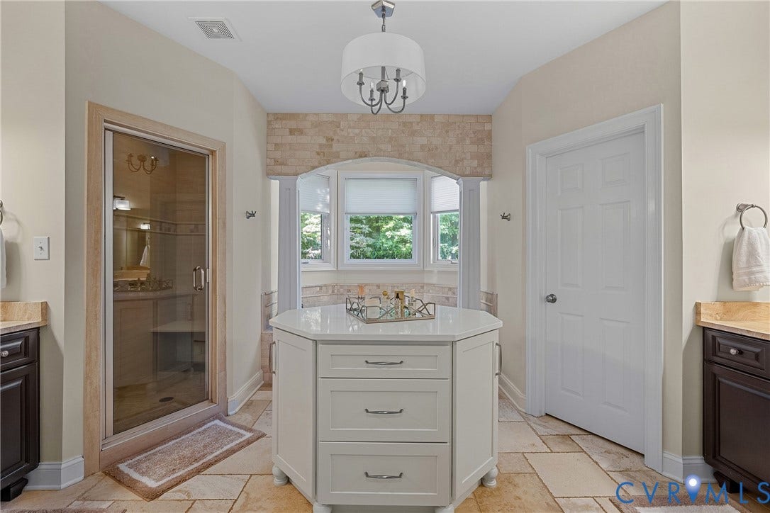 Walk-in shower with clear glass enclosure, central storage island with white countertop, bay window with upper stone-look tile accent, and light-toned tile flooring