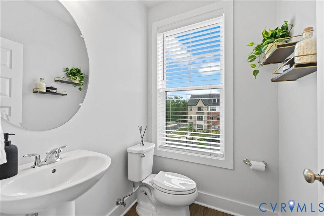 Pedestal sink with chrome faucet, circular wall mirror, window with blinds, and white toilet