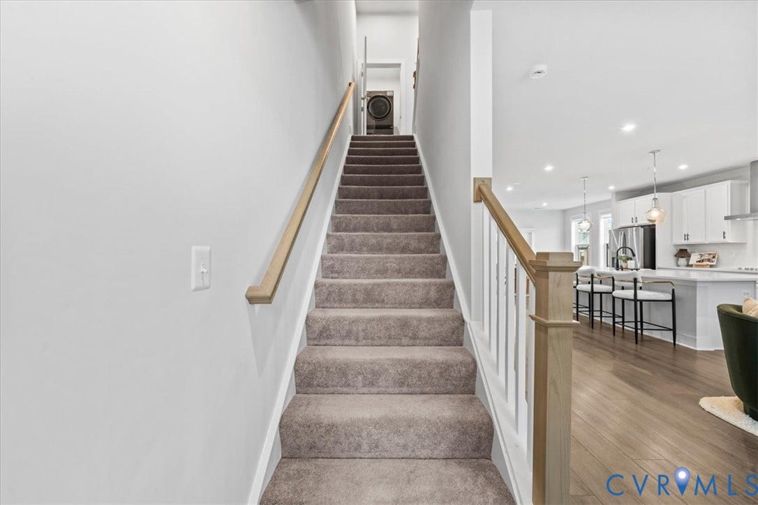 Carpeted staircase with wood handrails, bright open-concept living area with wood-finish flooring, white cabinetry, and stainless steel appliances
