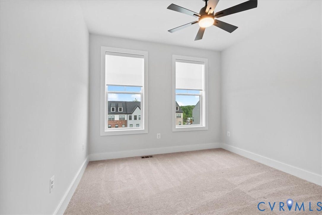 Neutral-toned carpeting, white walls, two windows with roller blinds, and a ceiling fan with integrated lighting