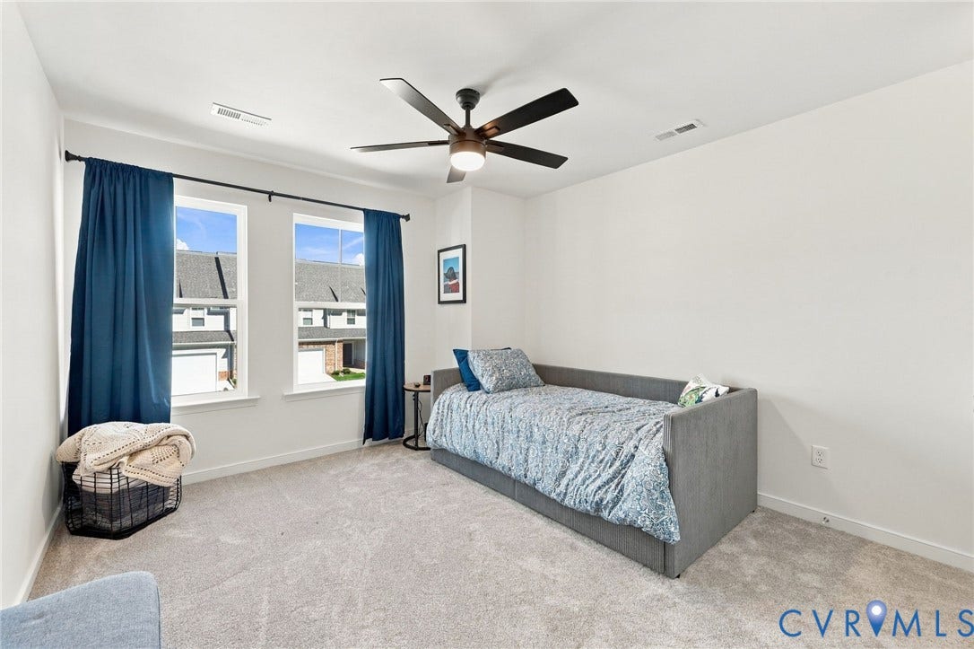 Bright room with light gray carpeting, two large windows, a ceiling fan with integrated lighting, and neutral wall tones