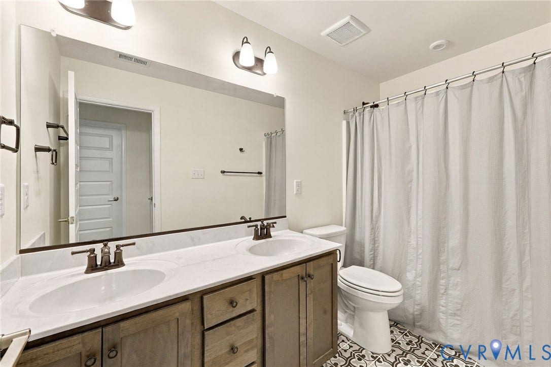 Double vanity with individual sinks, wood-finish cabinetry, patterned floor tiling, and wall-mounted light fixtures