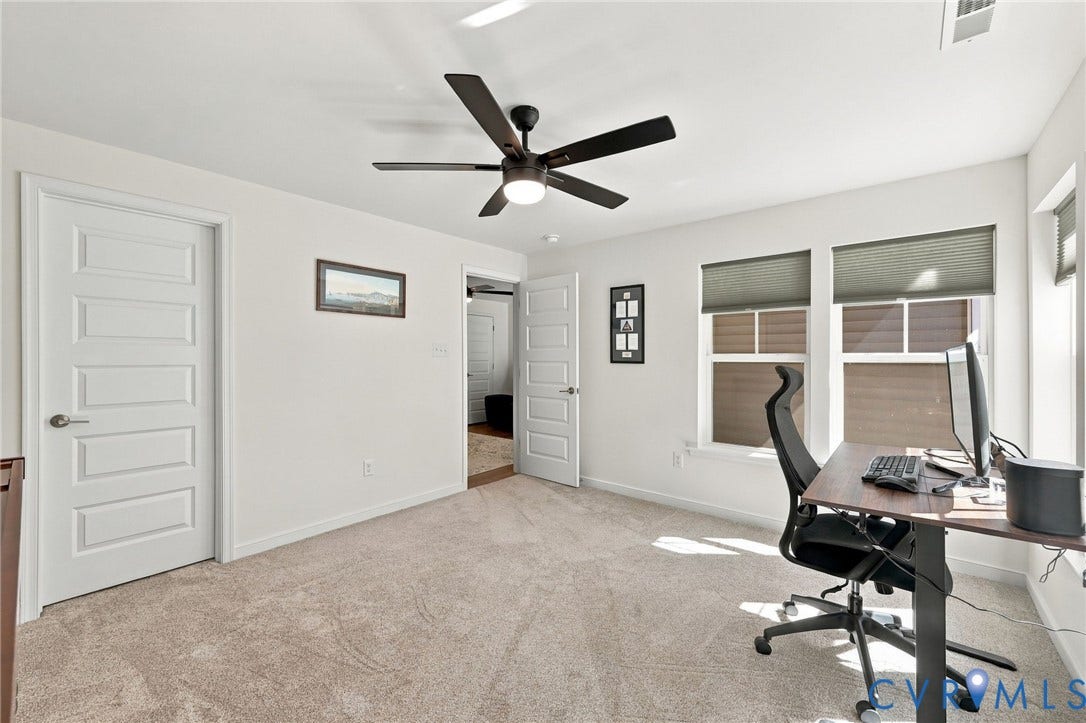 Plush carpeted flooring, a dark-toned ceiling fan with integrated lighting, multiple windows with cellular blinds, and white paneled doors