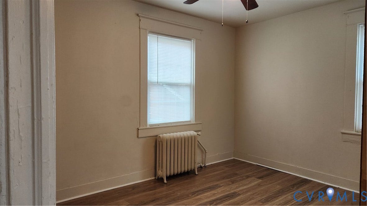 Wood-finish flooring, neutral wall tones, window with blinds, classic radiator, and a ceiling fan
