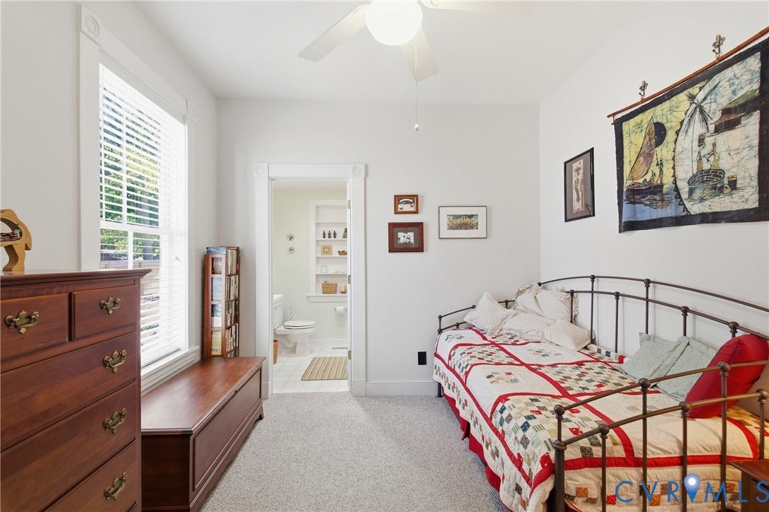 Carpeted room with a ceiling fan, a window with horizontal blinds, and a doorway revealing a bathroom with built-in shelving