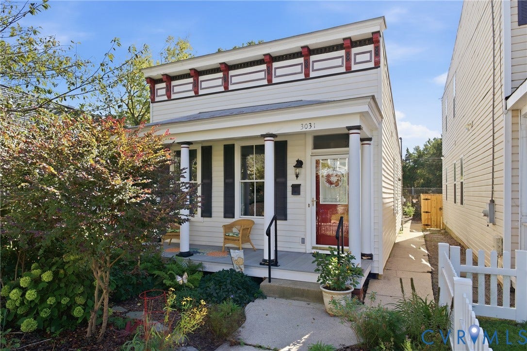 White siding exterior with decorative cornice detailing, a covered front porch, black window shutters, a red entry door, and a picket fence