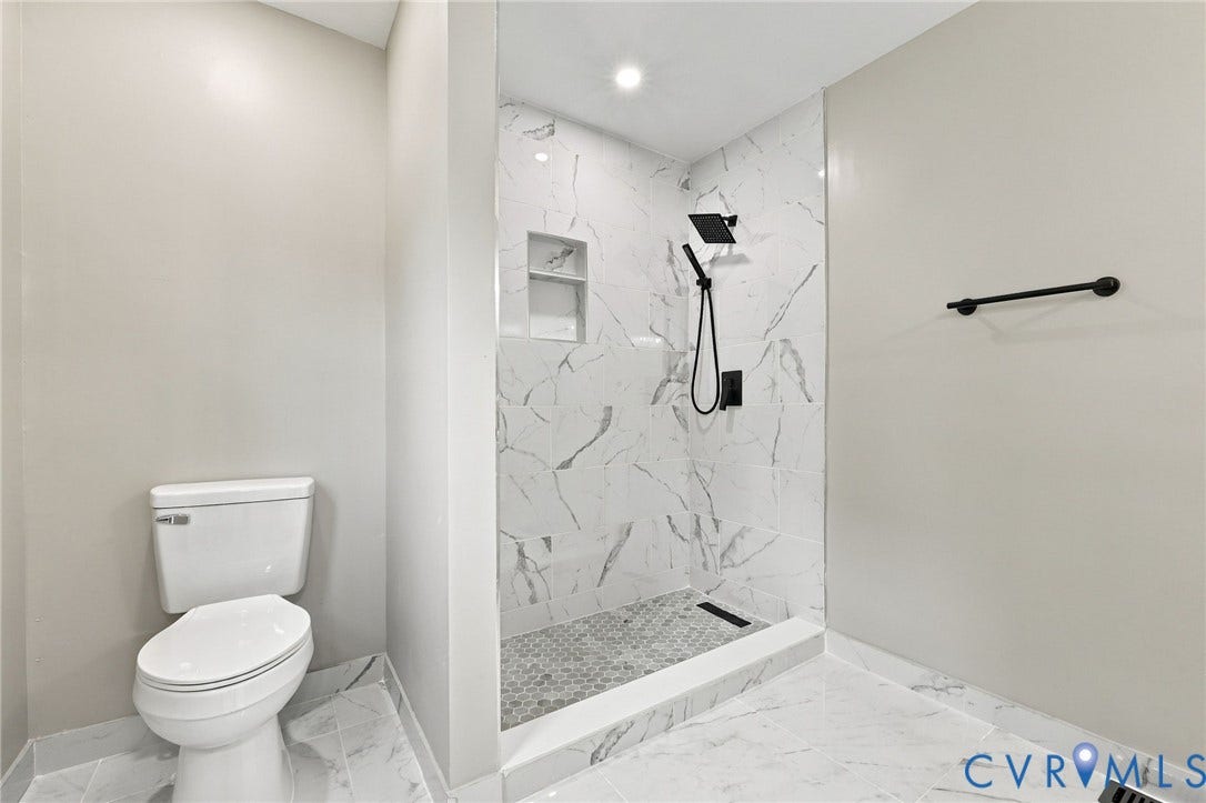 5831 Larrymore Road, Richmond, VA 23225 Walk-in shower featuring large-format stone-patterned tile, matte black rainfall showerhead with handheld sprayer, built-in recessed wall niche, and hexagon tile flooring, paired with a standard-height porcelain toilet and matte black wall-mounted towel bar