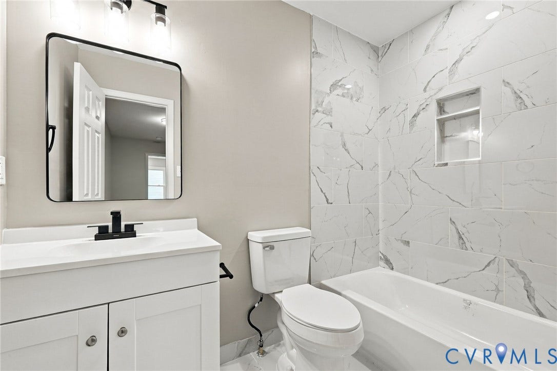 5831 Larrymore Road, Richmond, VA 23225 White vanity with a rectangular sink, matte black faucet, rectangular mirror with a black frame, and a bathtub surrounded by large-format, veined tile with a built-in recessed niche