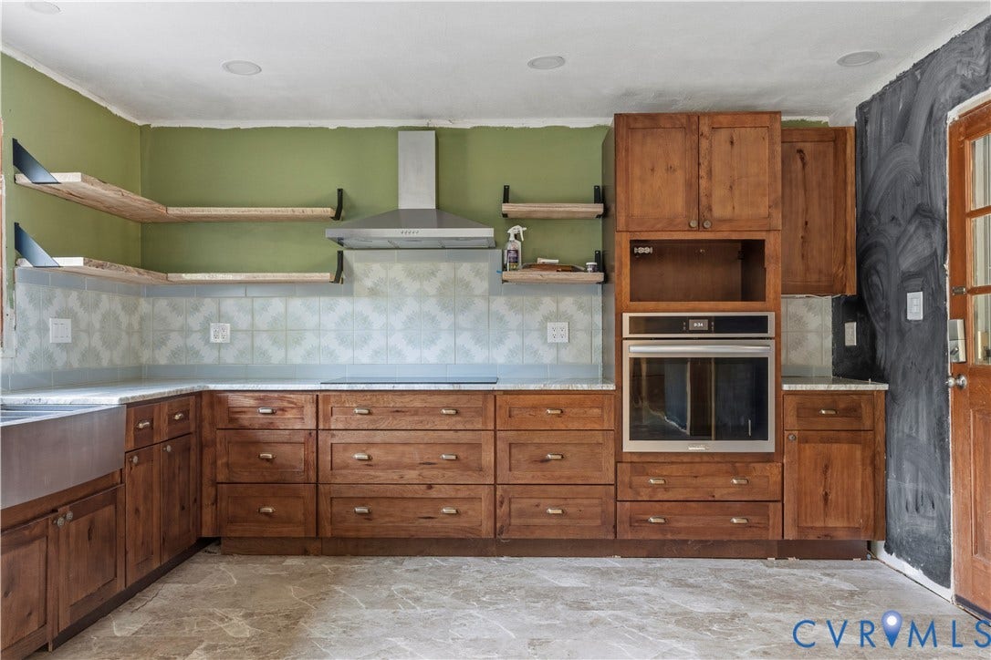 Wood cabinetry with brushed brass hardware, light-toned countertops, patterned tile backsplash, stainless steel range hood, and a built-in oven