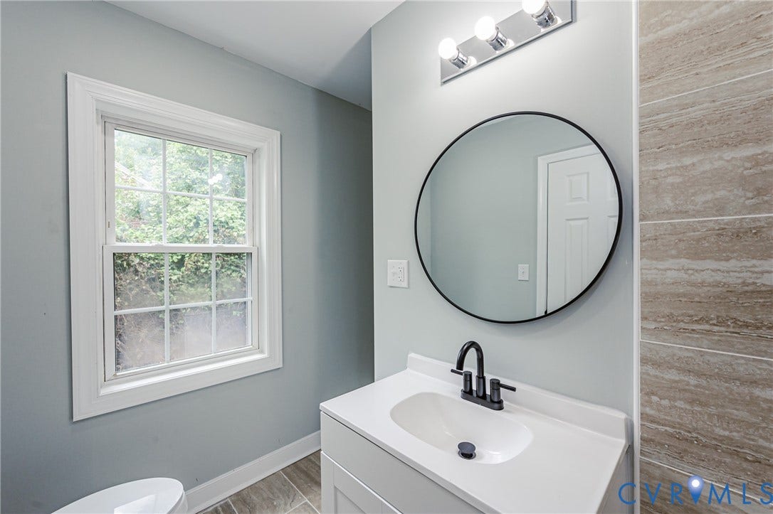 White framed window, light gray wall paint, tiled flooring, vanity with white countertop and integrated sink, black faucet and round mirror