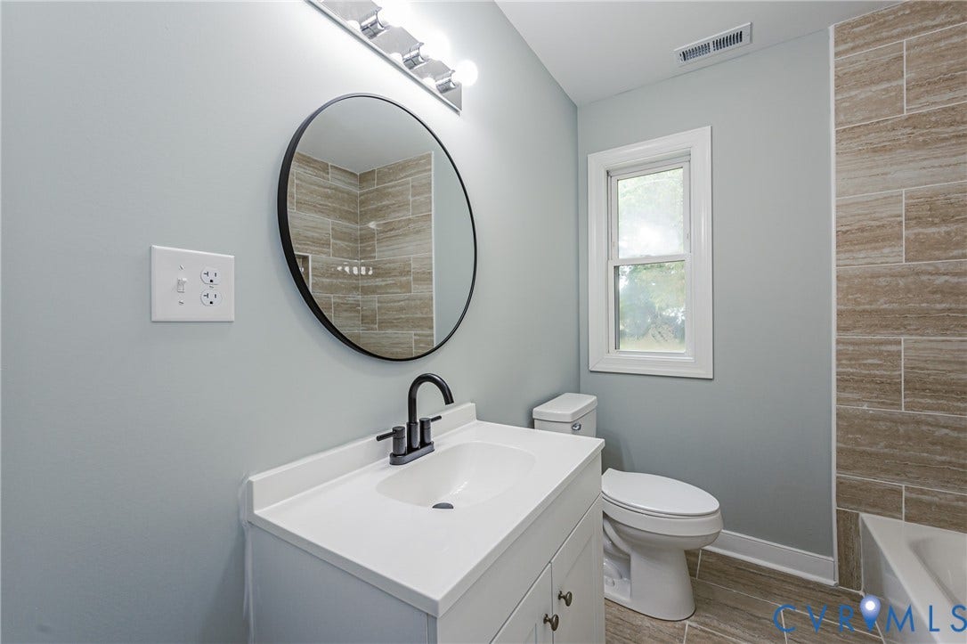 Vanity with integrated sink and matte black faucet, round wall mirror, bath with tile surround, and a toilet