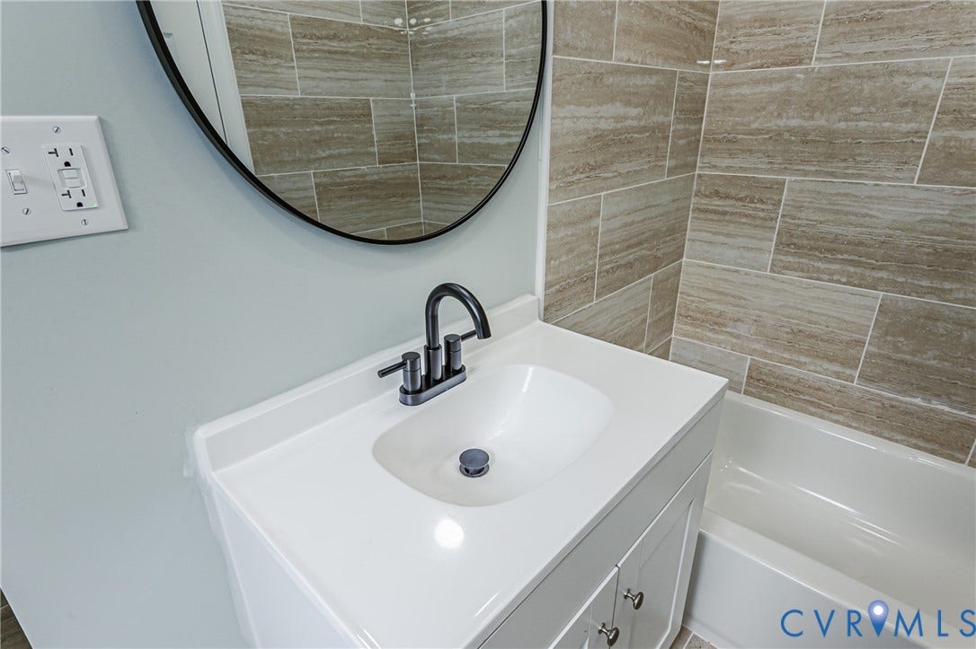 White vanity with integrated sink, matte black faucet, round framed mirror, and tiled shower surround