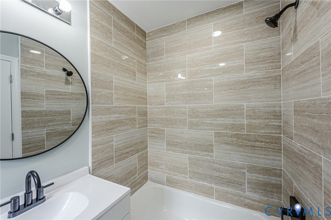 Full tile surround with horizontal accent pattern, white bathtub, black shower fixture, white vanity with integrated sink, and black-framed circular mirror