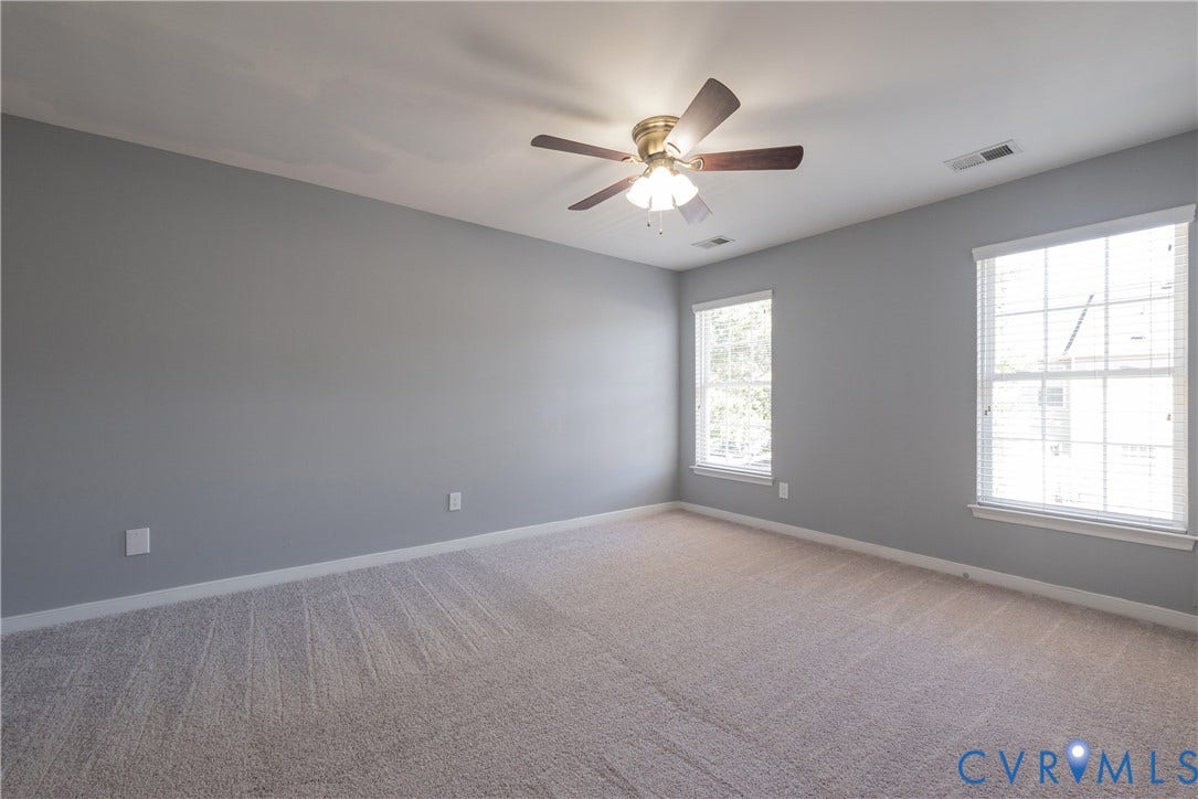 Spacious room featuring neutral carpeting, light gray wall paint, two windows with blinds, and a ceiling fan with integrated lighting