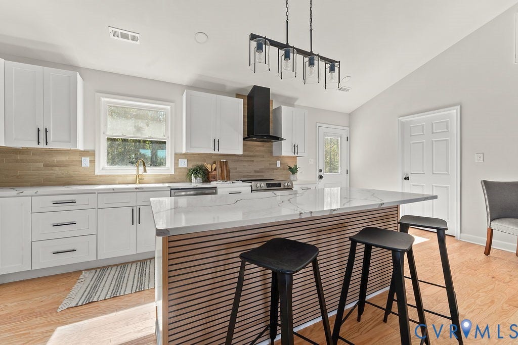 Vaulted ceiling with a linear pendant light fixture, white cabinetry with dark hardware, a stone countertop island, light wood-finish flooring, and a tiled backsplash