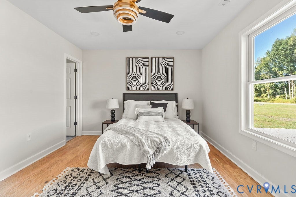 Bright, light-filled bedroom featuring hardwood flooring, a large picture window with natural landscape views, ceiling fan with brass and black finish, geometric wall art, and a patterned area rug anchoring the space