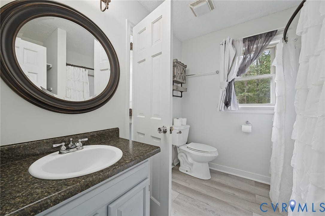 Bathroom featuring a vanity with a dark-toned countertop, integrated oval basin, oval wall-mounted mirror, and wood-finish flooring