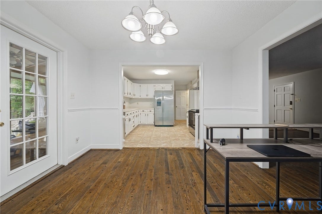 Dining area featuring dark wood-finish flooring, white chair rail molding, and a multi-arm chandelier, encompassing a full-light glass panel door and tiled kitchen with white cabinetry
