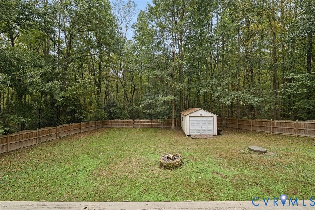 Fenced backyard featuring a detached storage building, stone fire pit, and mature tree line