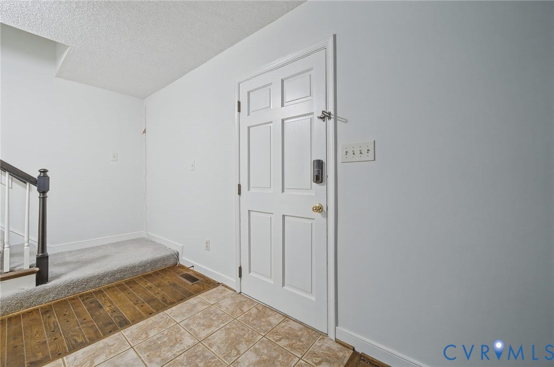 Six-panel white entry door with brass-tone hardware, wood-finish flooring, ceramic tile flooring, and staircase with a stained wood handrail and white balusters