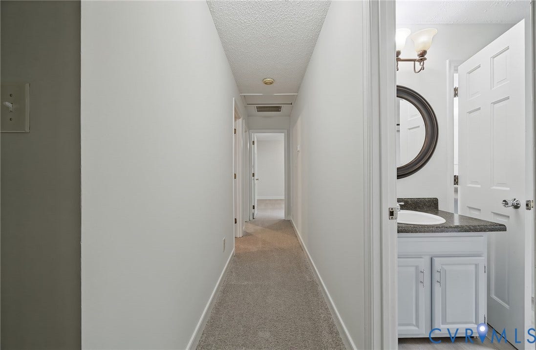 Hallway featuring carpeted flooring and a multi-panel door leading to a bathroom equipped with a white vanity, solid surface countertop, drop-in sink, and a circular wall mirror