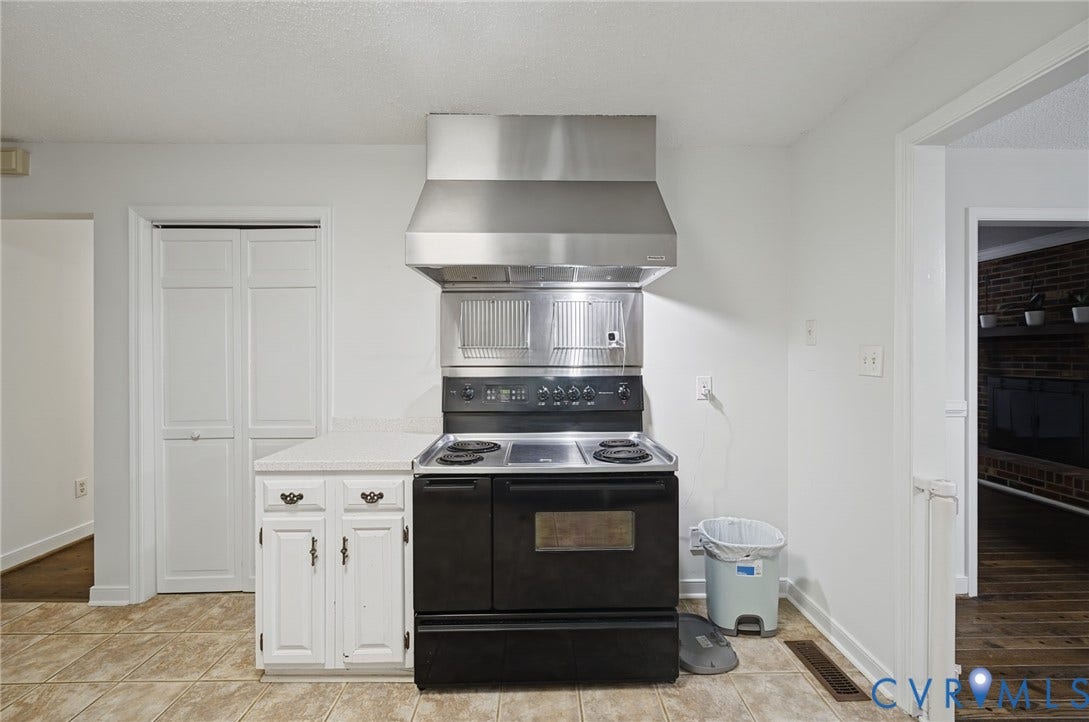 Stainless steel range hood, black electric range, white cabinetry with countertop, and tile flooring