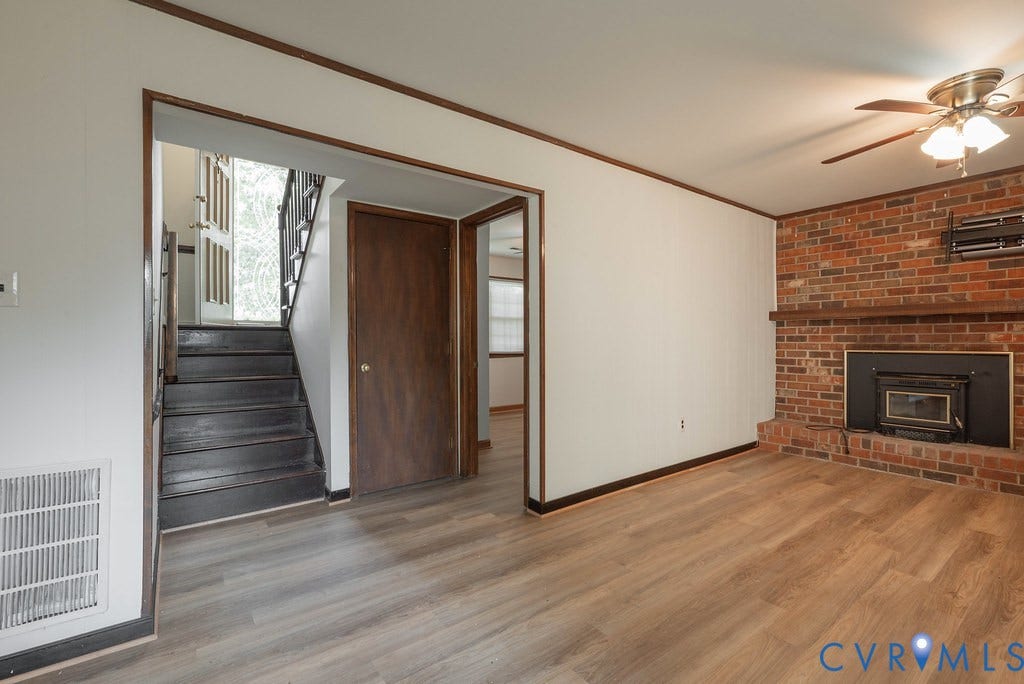 Wood-finish flooring, brick fireplace with a wood mantel, ceiling fan with integrated lighting, and a dark wood staircase with a white paneled door