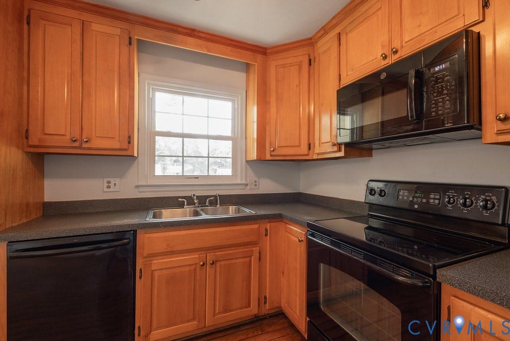 Wood-finish cabinetry with brass hardware, black countertops, stainless steel double basin sink, black dishwasher, and black range with an overhead microwave