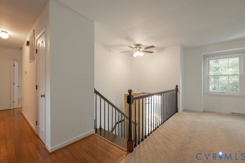 Open landing area featuring carpeted flooring, a ceiling fan with integrated lighting, a large window, a wood-finish handrail with dark balusters, and a hallway with wood-finish flooring