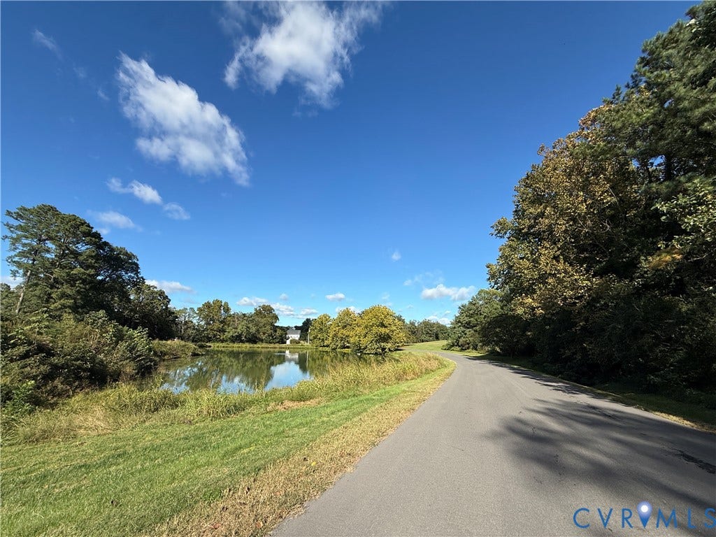 #30 Molly's Way, Urbanna, VA 23175 Paved road with flanking green spaces, mature trees, and a reflective pond