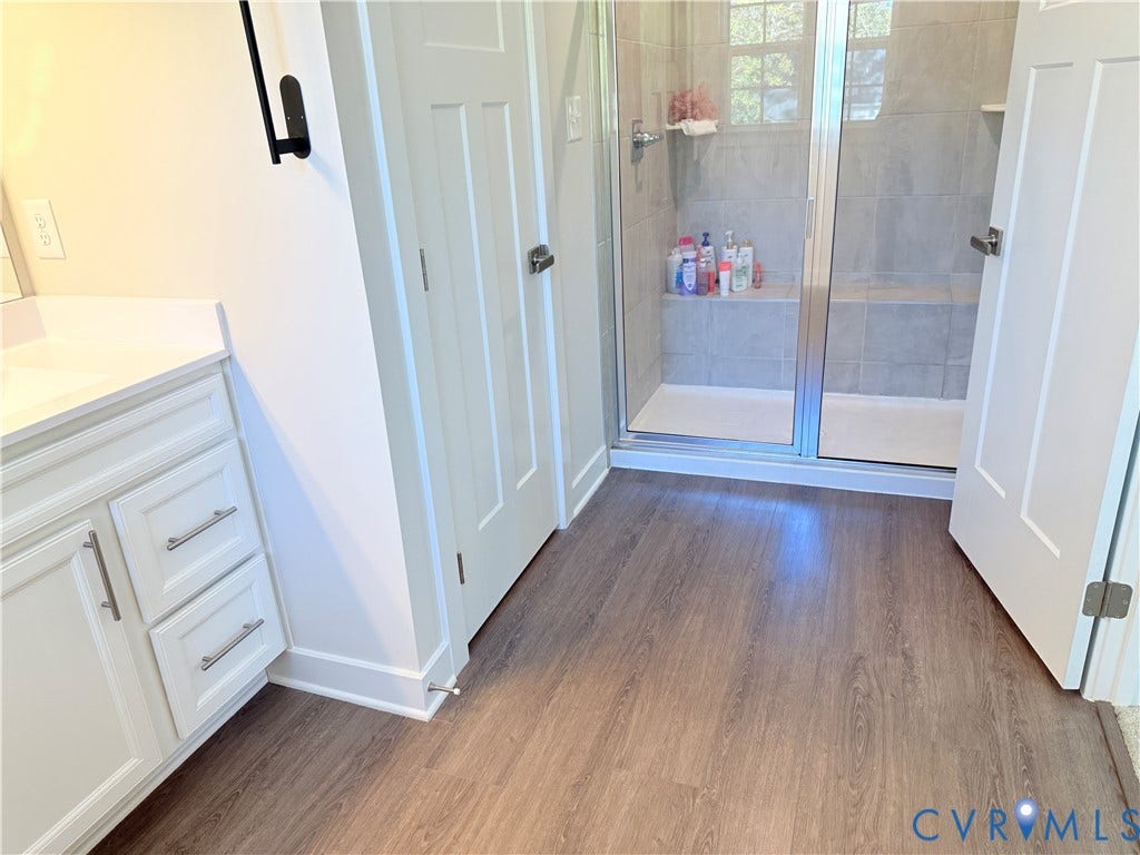 14225 Botsford Avenue, Wellspring, Chester, VA 23831 White vanity with brushed metal hardware, walk-in shower with a built-in bench, neutral tile shower surround, glass shower enclosure, and wood-finish flooring
