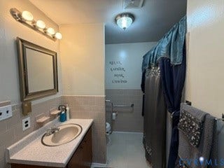 Spa-inspired bathroom featuring a round vessel sink with polished chrome faucet, quartz countertop, illuminated vanity mirror with integrated lighting, tile wainscoting, and a separate shower enclosure with dark tile surround and coordinated towel bars