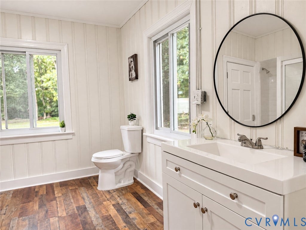 Wood-finish flooring, white paneled walls, a single vanity with an integrated sink, a circular mirror, and a two-panel window