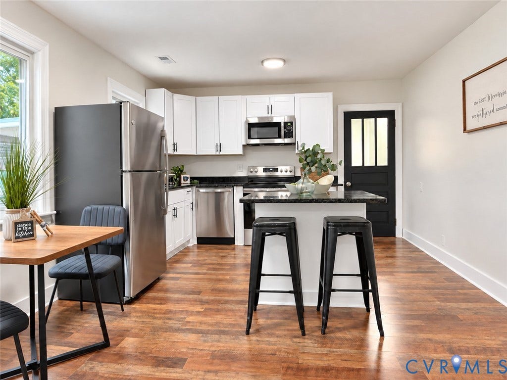 Kitchen featuring wood-finish flooring, white cabinetry, stainless steel appliances, a center island with a dark countertop, and a black paneled door