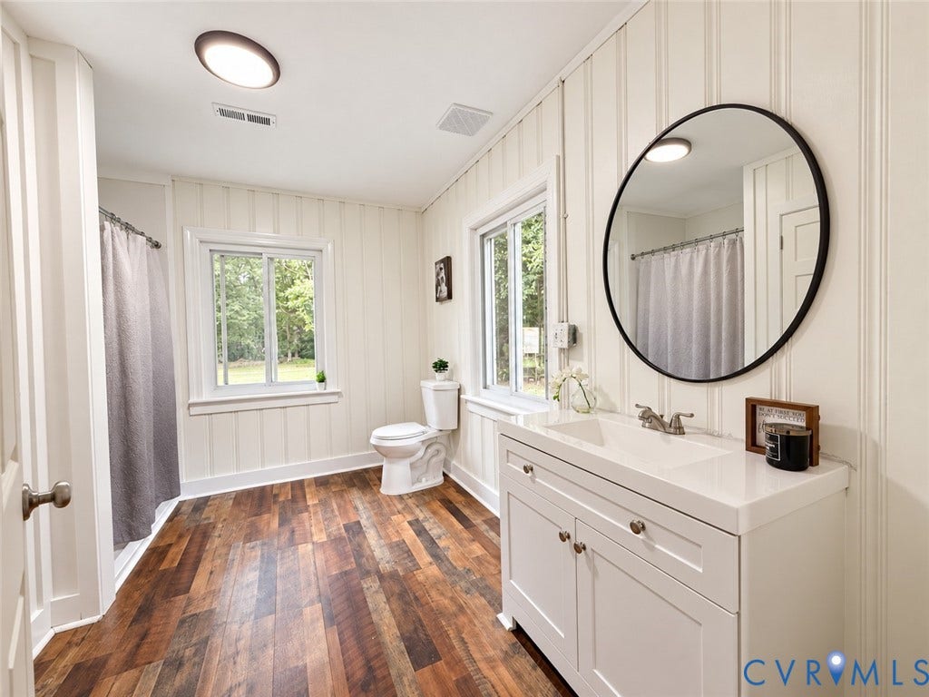 White beadboard wall paneling, multi-tone wood-finish flooring, white vanity with integrated sink, chrome faucet, and a round black-framed mirror