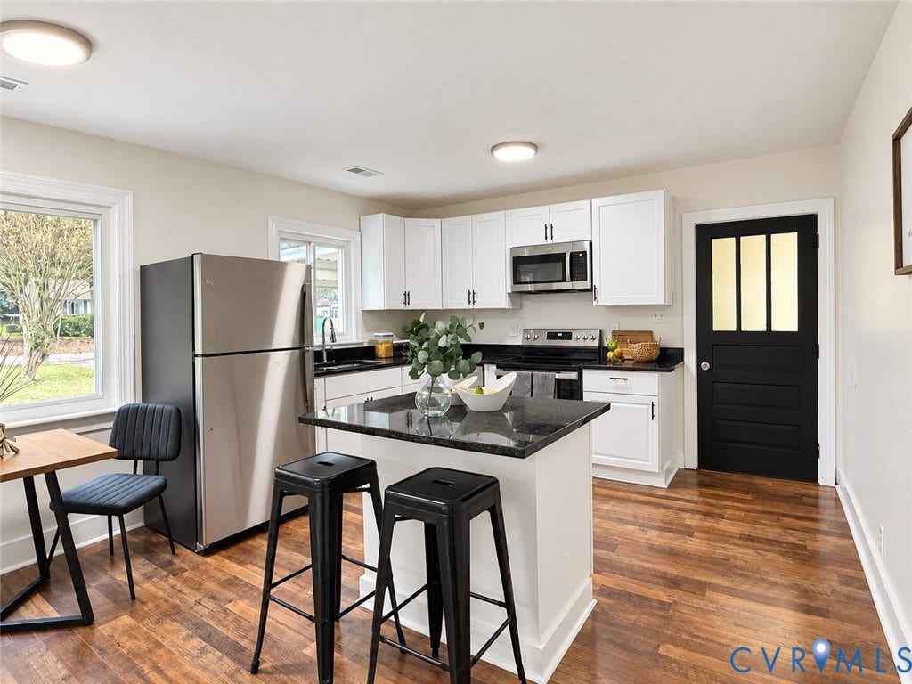 Kitchen featuring wood-finish flooring, a central island with a dark countertop, white cabinetry with dark hardware, stainless steel appliances, and a black paneled door