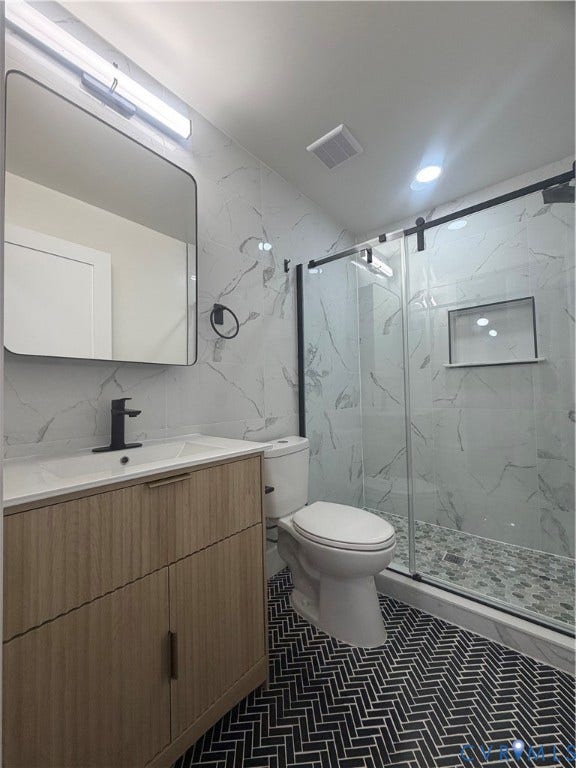 Modern bathroom featuring a wood-finish vanity with an integrated sink, black herringbone tile flooring, and a glass-enclosed shower with a recessed shelf and pebble tile floor