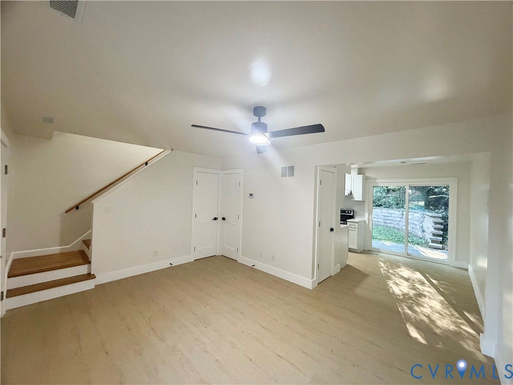 Light wood-finish flooring, white walls, black ceiling fan with integrated lighting, wood handrail on staircase, and sliding glass door