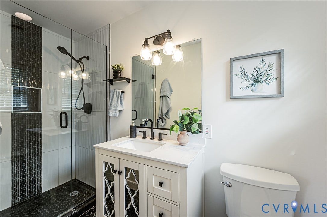 1608 Lochwood Drive, Henrico, VA 23238 Spa-inspired bathroom featuring a frameless glass-enclosed shower with mosaic tile accents, a white vanity with geometric cabinet detailing and quartz countertop, dual sconces with Edison-style bulbs, and a low-flow toilet