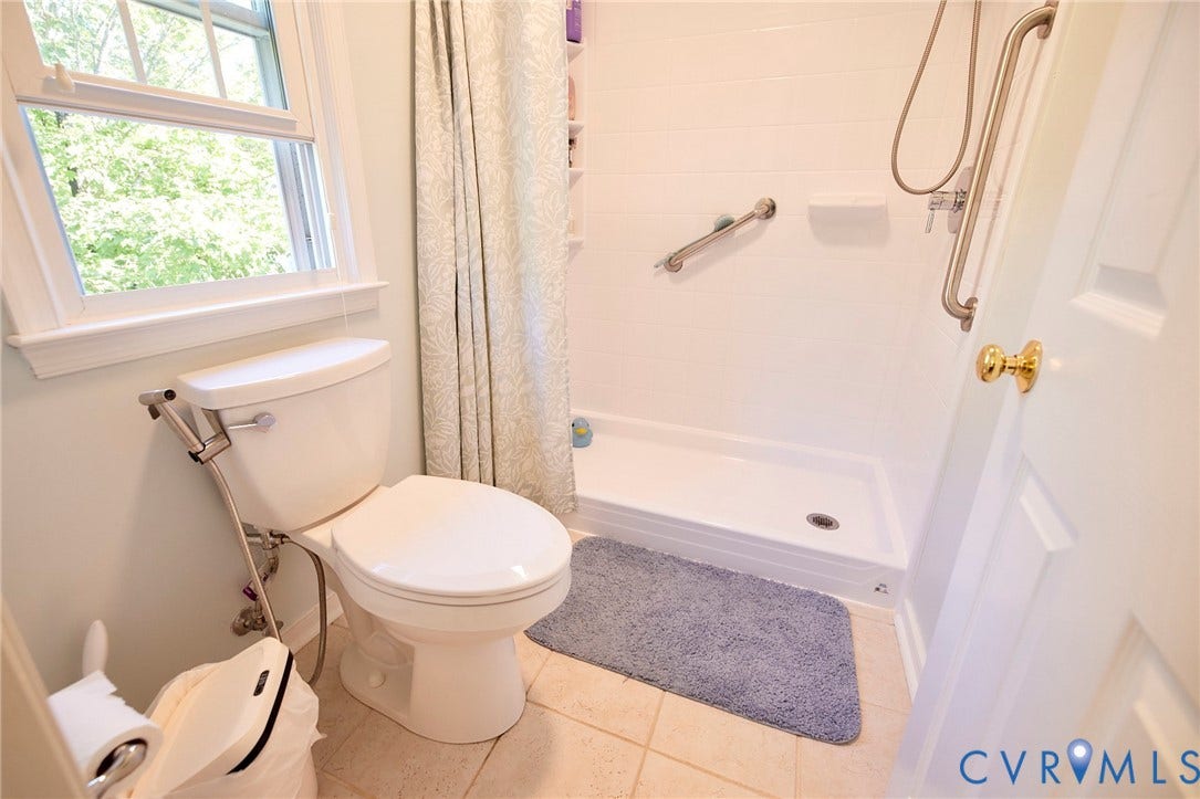 2633 Bradway Lane, Henrico, VA 23233 Bright, updated bathroom featuring a white ceramic toilet with dual flush mechanism, tile flooring, a walk-in shower enclosure with glass door and grab bars, a window with natural light, and brushed nickel hardware throughout