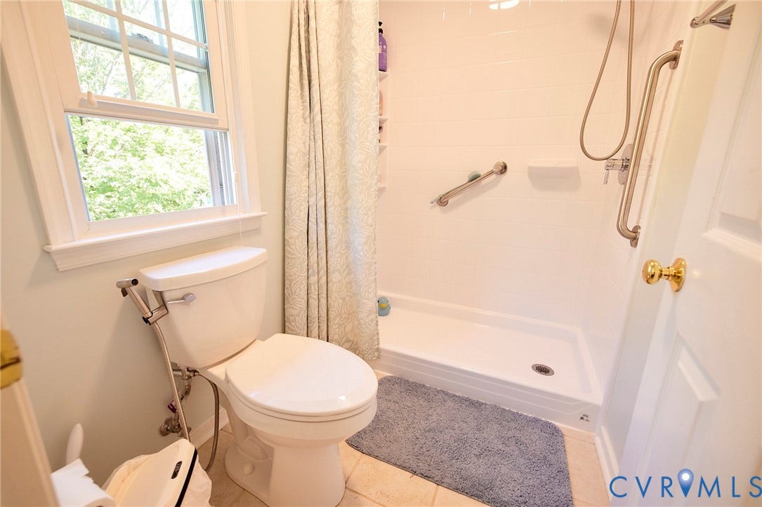 2633 Bradway Lane, Henrico, VA 23233 Clean, updated bathroom featuring a porcelain toilet with dual-flush mechanism, a tile-surround shower with glass enclosure and chrome fixtures, ceramic tile flooring, and a large window providing natural light