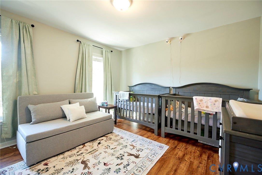 2633 Bradway Lane, Henrico, VA 23233 Bright nursery featuring hardwood flooring, two dark wood cribs with slatted design, large windows with soft green drapery providing natural light, and a comfortable upholstered sofa ideal for seating or lounging