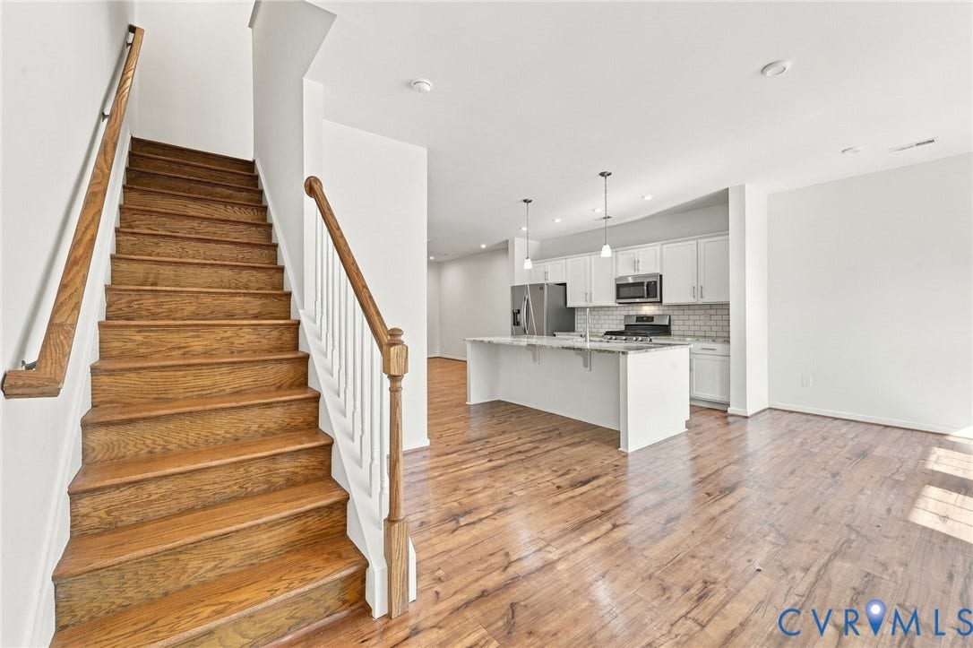 10818 Ashton Poole Place, Henrico, VA 23059 Open-concept living space featuring rich wood-finish flooring throughout, a modern kitchen with white cabinetry, stainless steel appliances, and a tile backsplash, complemented by a grand wooden staircase with white railings and natural wood treads