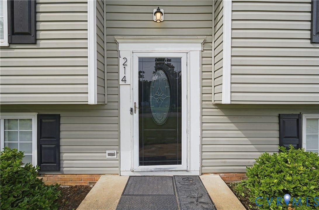 Light-colored horizontal siding, a storm door with an oval decorative glass insert, a white entry door frame, and black exterior shutters