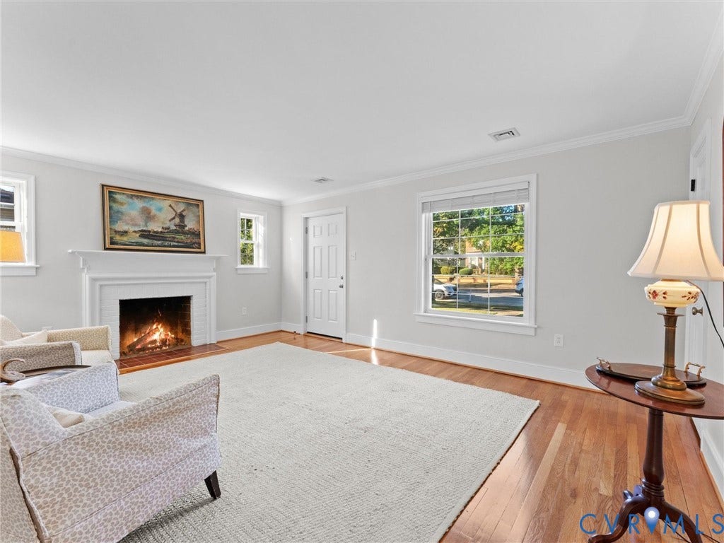 Hardwood flooring throughout, white painted brick fireplace with mantel, white trim, multiple windows with natural light, and a paneled entry door