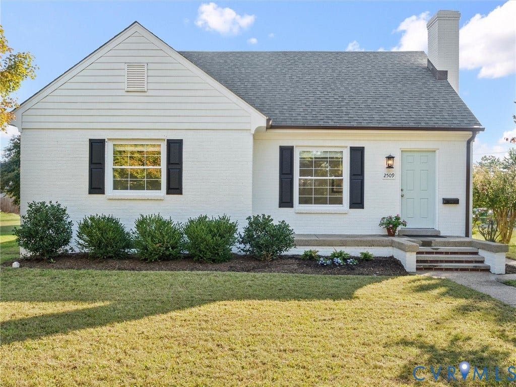 White brick exterior with a light blue entry door, black window shutters, a prominent chimney, and mature foundation plantings