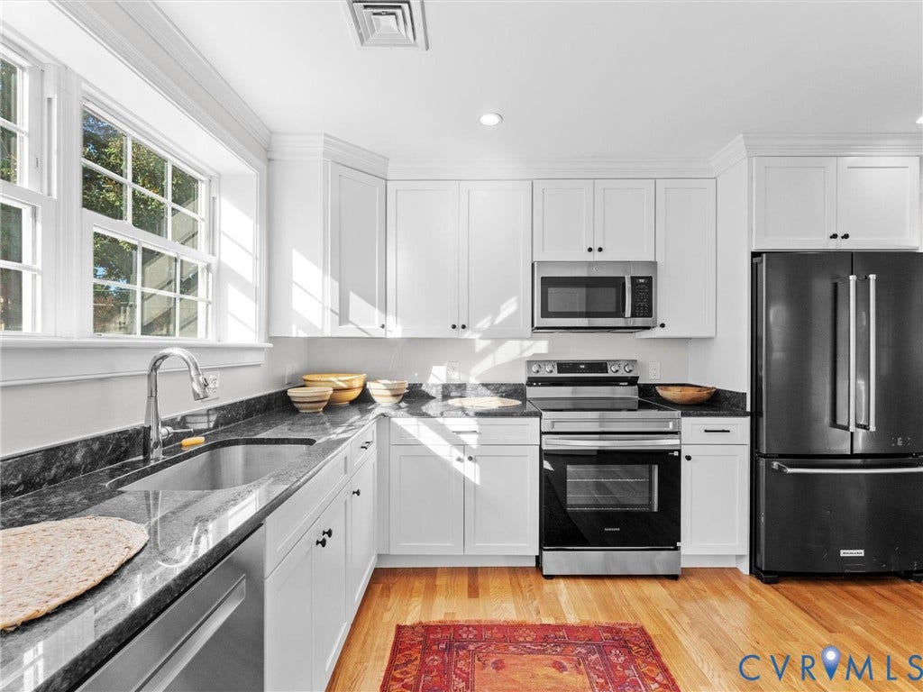 White cabinetry with dark contrasting hardware, stainless steel appliances, dark stone countertops, wood-finish flooring, and a large window with muntins