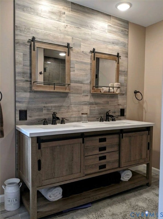 Dual vanity with a white countertop and integrated sinks, wood-finish cabinetry, matte black fixtures, and a tiled accent wall